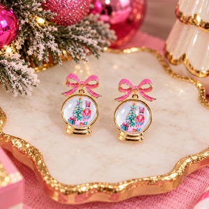 May include: A pair of gold-toned earrings shaped like snow globes, each topped with a glittery pink bow. Inside the clear globes are miniature illustrations of a nutcracker, a decorated Christmas tree, and colorful gift boxes.