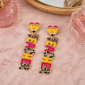May include: A pair of dangling earrings with a yellow pencil-shaped top and the word "TEACHER" in pink and leopard print. The earrings are set against a pink background with a gold-colored mirror and perfume bottles.
