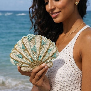 May include: A beaded clutch in a fan shape, featuring a design of white, teal, and gold beads. The clutch is held against a backdrop of the ocean, suggesting a beach or resort theme. The clutch has a gold beaded border.