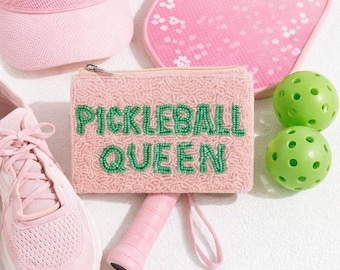 May include: A collection of pink pickleball gear, including a visor, paddle, beaded pouch with "PICKLEBALL QUEEN" text, sneakers, and green balls. The paddle has a pink handle. The paddle has a pink handle.