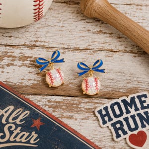 May include: A pair of earrings featuring a gold-tone metal bow in bright blue enamel attached to a round, white, textured baseball charm with red stitching details. The earrings are displayed alongside a baseball, a wooden bat, and a sign.