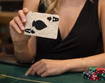 Puede incluir: Clutch con cuentas de color crema con un diseño de As de Picas negro. El bolso tiene una cremallera dorada y detalles negros. La imagen está ambientada sobre un fondo de mesa de casino verde.