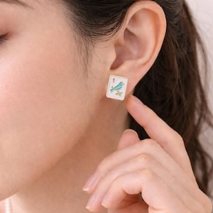 May include: Square gold-tone stud earrings. Each earring features a white background with a teal bird and green leaves. The number "1" is in the upper left corner. The earrings are on a person's ear.
