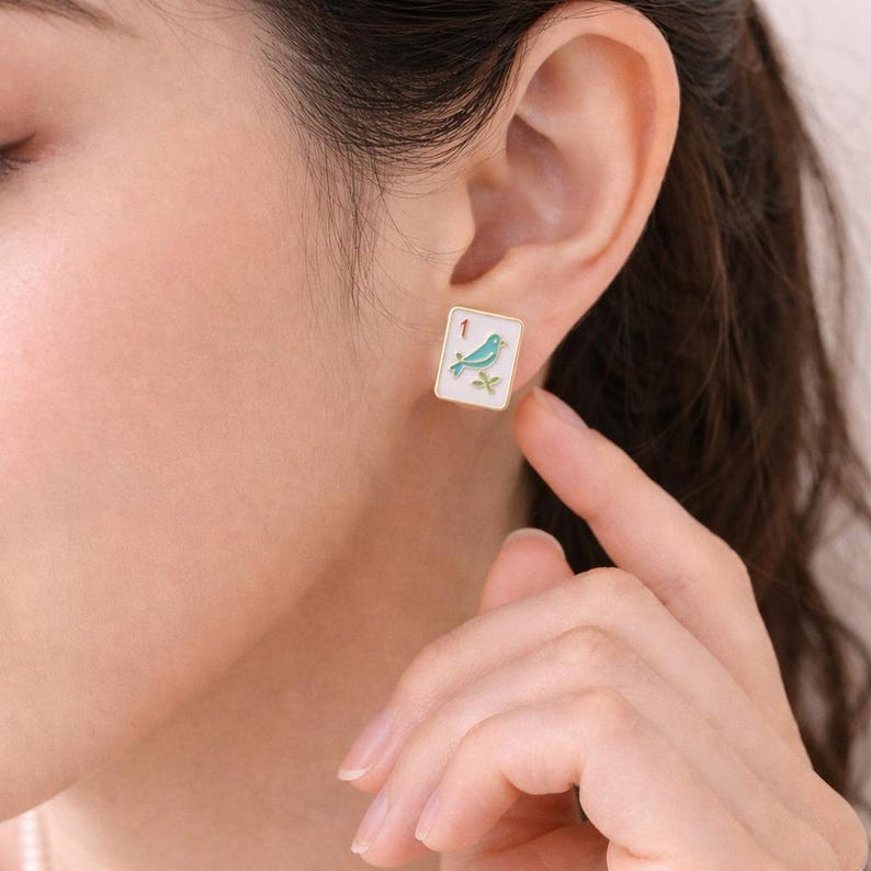 May include: Square gold-tone stud earrings. Each earring features a white background with a teal bird and green leaves. The number "1" is in the upper left corner. The earrings are on a person's ear.