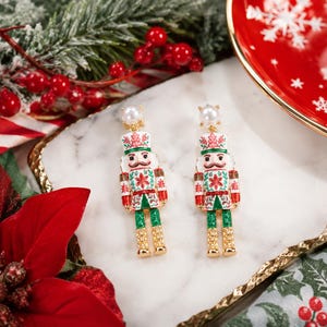 May include: A pair of dangle earrings featuring nutcracker figures with white, red, and green enamel detailing, gold-tone accents, and a small white faux pearl at the top of each earring post, designed with a festive holiday aesthetic.