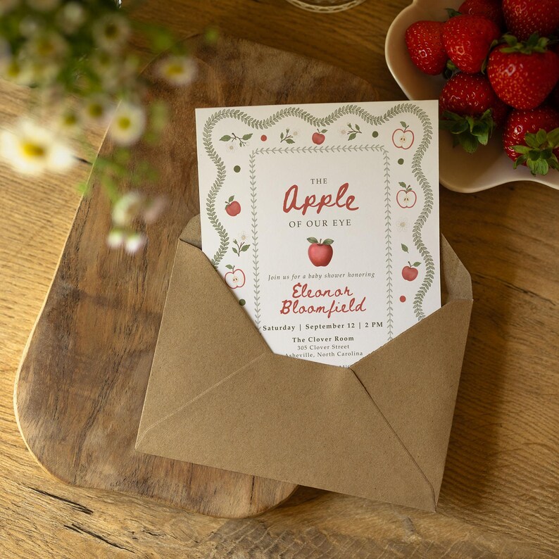 May include: A white rectangular baby shower invitation card tucked into a brown kraft paper envelope. The card features a green leaf border, red apple illustrations, and text reading The Apple of Our Eye, honoring Eleanor Bloomfield.