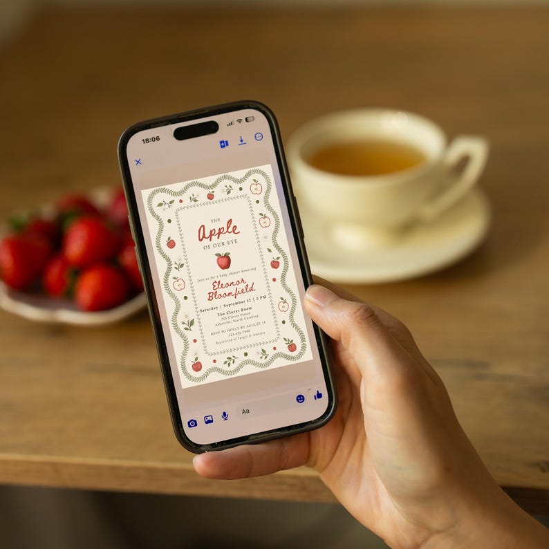 May include: A smartphone displaying a digital baby shower invitation with a cream background, red apple illustrations, and green leaf borders. The text reads: The Apple of Our Eye, honoring Eleanor Bloonfield, Saturday, September 12, 3 PM, The Clover Room.
