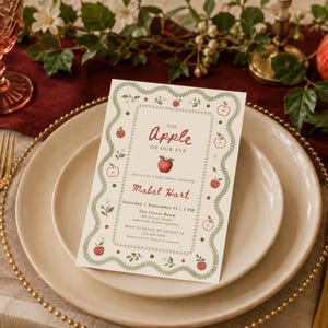 May include: A rectangular baby shower invitation card with a cream background, featuring a green leafy border and red apple illustrations. The text reads: The Apple of our eye, join us for a baby shower honoring Mabel Hart, Saturday, September 12, 2 PM.