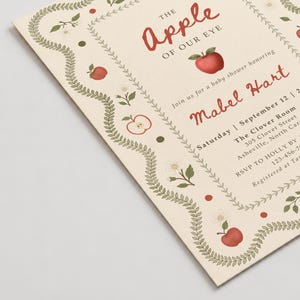 May include: A cream-colored baby shower invitation card featuring a decorative border of green leaves, white flowers, and red apples. The text reads: The Apple of Our Eye, Join us for a baby shower honoring Mabel Hart, Saturday, September 12.