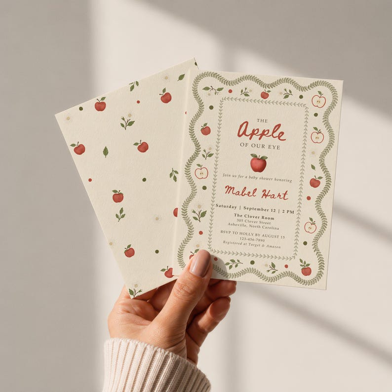 May include: A cream-colored baby shower invitation and matching envelope featuring a red apple pattern and green leaf border. The text reads: The Apple of Our Eye, join us for a baby shower honoring Mabel Hart, Saturday, September 12, 2 PM.