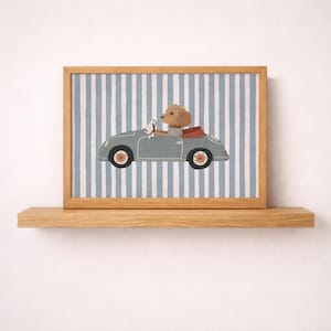 Könnte beinhalten: Ein gerahmter Druck, der einen hellbraunen Hund am Steuer eines grauen Oldtimer-Cabriolets zeigt. Der Hintergrund besteht aus vertikalen hellblauen und weißen Streifen. Der Rahmen aus hellem Holz steht auf einem Holzregal.