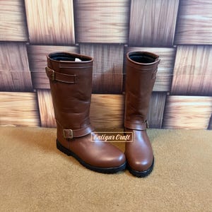 May include: A pair of mid-calf brown boots featuring two adjustable straps with brass-toned buckles, one at the ankle and one at the top. The boots have a smooth finish, a rounded toe, and a black sole. The text Caligar Craft is visible.
