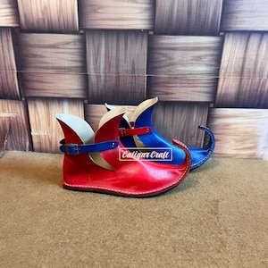 May include: A pair of pointed-toe shoes featuring a curved, upturned front. One shoe is red with a blue strap and buckle, while the other is blue with a red strap and buckle. The text Caligar Craft is visible on the side of the red shoe.