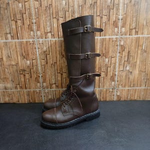 Puede incluir: Botas altas de cuero marrón con cordones en el tobillo y tres correas con hebilla. Las botas tienen suelas negras y están hechas de cuero liso y duradero. Altura aproximada de 40 cm.