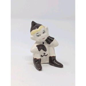 May include: A small ceramic figurine of a seated elf with a glossy white finish, featuring dark brown painted details on the pointed hat, gloves, boots, and facial features. The elf has a cream-colored band on the hat and a small dark mark on the chest.