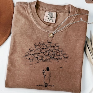 Op de afbeelding: Espresso-kleurig Comfort Colors T-shirt met een ronde hals. Het shirt heeft een afbeelding van een persoon die een schaap vasthoudt, met een kudde schapen op de achtergrond. Een zilveren ketting met een hanger ligt op het shirt.