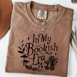 May include: Espresso-colored t-shirt with the text "In My Bookish Era" in a whimsical font, featuring mushroom and leaf illustrations. A silver necklace with a pendant rests on the shirt. The shirt is a Comfort Colors brand.