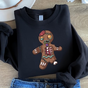 May include: A black long-sleeve sweatshirt featuring a graphic of a brown gingerbread person designed as a zombie. The character has exposed brain matter, glowing yellow eyes, green drool, and a rib cage visible through a torn chest area.