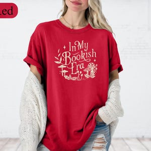 May include: Red t-shirt with the text "In My Bookish Era" in a circular design with botanical and mushroom illustrations. The shirt is a solid red color and has short sleeves.