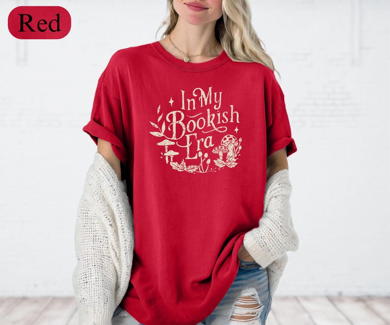 May include: Red t-shirt with the text "In My Bookish Era" in a circular design with botanical and mushroom illustrations. The shirt is a solid red color and has short sleeves.