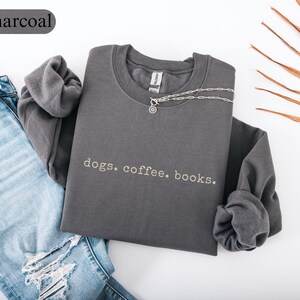 May include: Charcoal-colored sweatshirt with the text "dogs. coffee. books." in white. The long-sleeved shirt is folded, with a silver necklace draped across it. A pair of blue jeans is visible on the left side of the image.