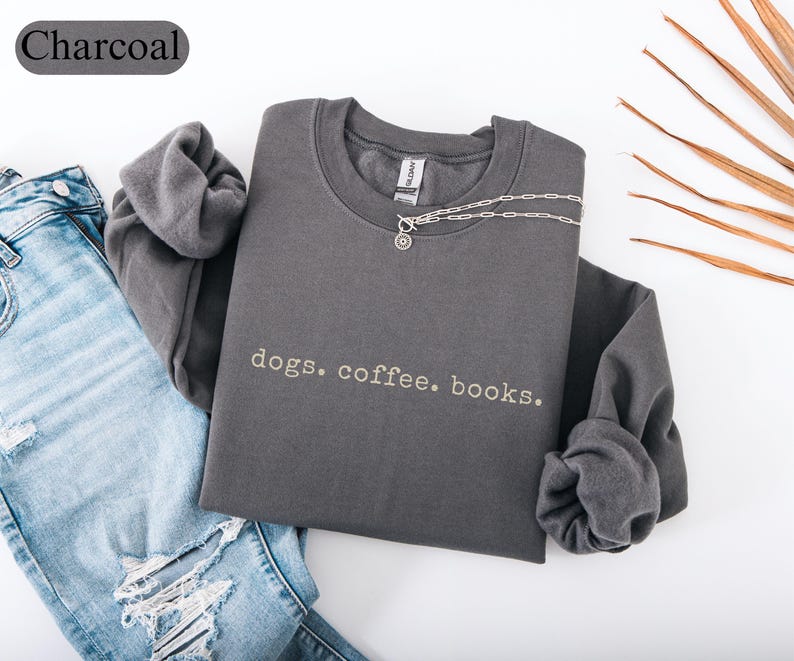 May include: Charcoal-colored sweatshirt with the text "dogs. coffee. books." in white. The long-sleeved shirt is folded, with a silver necklace draped across it. A pair of blue jeans is visible on the left side of the image.