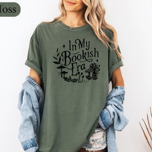 May include: Moss green t-shirt with the text "In My Bookish Era" in a black, circular design with mushroom and leaf illustrations. The shirt has short sleeves and a relaxed fit.