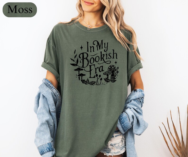 May include: Moss green t-shirt with the text "In My Bookish Era" in a black, circular design with mushroom and leaf illustrations. The shirt has short sleeves and a relaxed fit.
