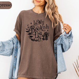 May include: Espresso-colored t-shirt with the text "In My Bookish Era" in a black, whimsical font. The design includes illustrations of mushrooms and foliage. The shirt is paired with a light blue denim jacket.