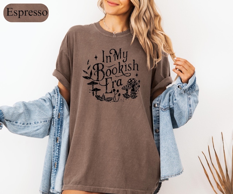 May include: Espresso-colored t-shirt with the text "In My Bookish Era" in a black, whimsical font. The design includes illustrations of mushrooms and foliage. The shirt is paired with a light blue denim jacket.