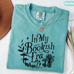 May include: Seafoam green t-shirt with the text "In My Bookish Era" in black, surrounded by a botanical design. A gold necklace with a pendant rests on the shirt. The shirt is a Comfort Colors brand.