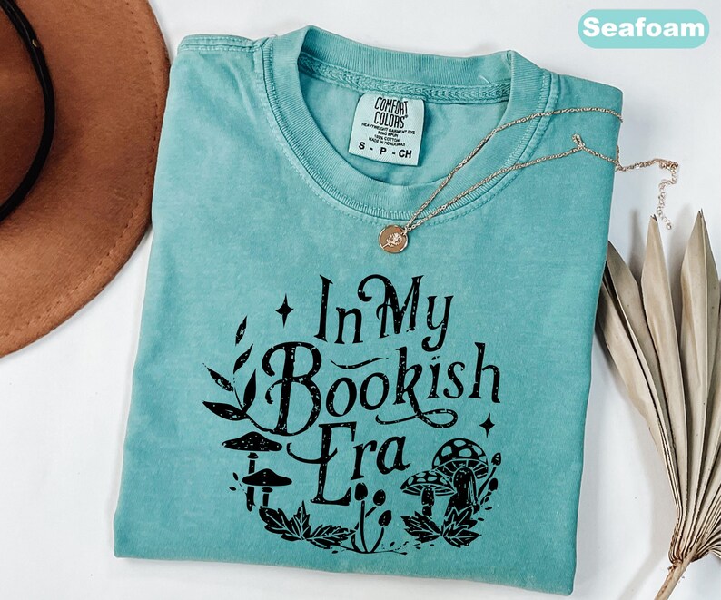 May include: Seafoam green t-shirt with the text "In My Bookish Era" in black, surrounded by a botanical design. A gold necklace with a pendant rests on the shirt. The shirt is a Comfort Colors brand.
