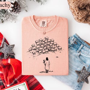 Op de afbeelding: Perzikkleurig T-shirt met ronde hals met een zwarte illustratie van een persoon die een schaap vasthoudt, met een kudde schapen op de achtergrond. Het woord "Peachy" staat in de linkerbovenhoek.