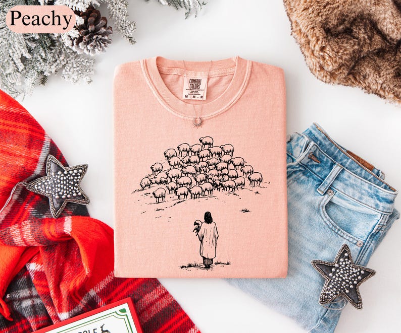Op de afbeelding: Perzikkleurig T-shirt met ronde hals met een zwarte illustratie van een persoon die een schaap vasthoudt, met een kudde schapen op de achtergrond. Het woord "Peachy" staat in de linkerbovenhoek.