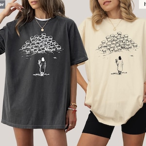 Op de afbeelding: Twee oversized T-shirts, &eacute;&eacute;n in peperkleur en &eacute;&eacute;n in ivoor, met een zwart-witte afbeelding. De afbeelding toont een persoon met een kudde schapen. De shirts hebben korte mouwen en een relaxte pasvorm.