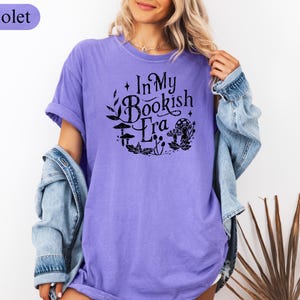May include: A violet-colored t-shirt with the text "In My Bookish Era" in a black, circular design featuring leaves, mushrooms, and other botanical elements. The shirt has short sleeves and a relaxed fit.