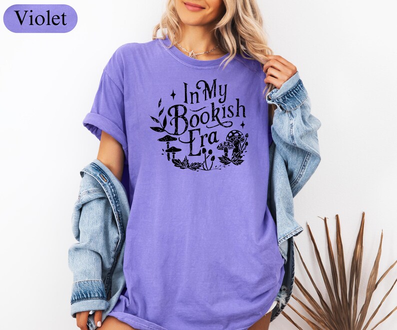 May include: A violet-colored t-shirt with the text "In My Bookish Era" in a black, circular design featuring leaves, mushrooms, and other botanical elements. The shirt has short sleeves and a relaxed fit.