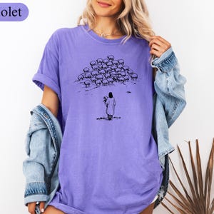 Op de afbeelding: Violetkleurig T-shirt met een zwart-wit illustratie van een persoon voor een kudde schapen. De persoon houdt een bundel vast. Het woord "Violet" staat in een violette doos linksboven.