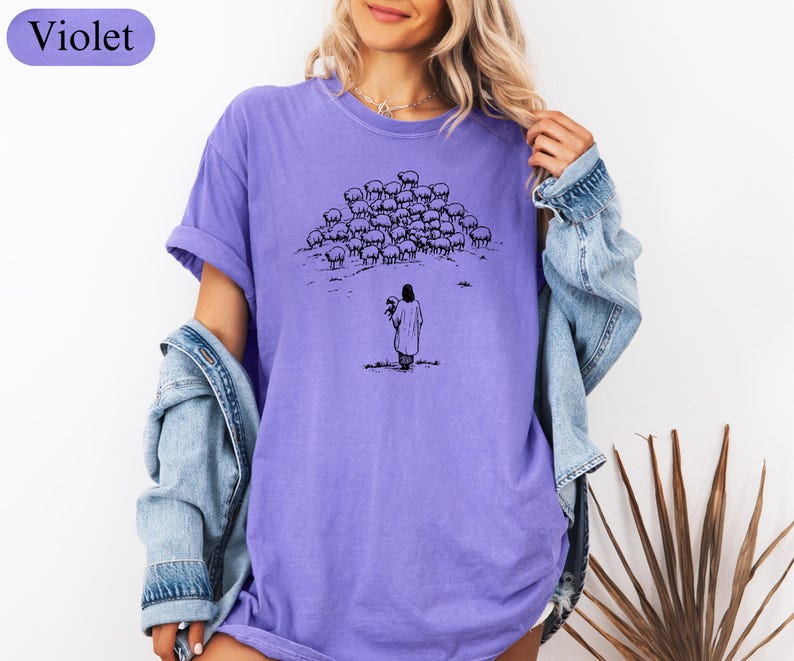 Op de afbeelding: Violetkleurig T-shirt met een zwart-wit illustratie van een persoon voor een kudde schapen. De persoon houdt een bundel vast. Het woord "Violet" staat in een violette doos linksboven.