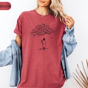 Op de afbeelding: Karmizijnrood T-shirt met een zwarte illustratie van een persoon en een kudde schapen. De persoon houdt een schaap vast. Het shirt heeft korte mouwen en een relaxte pasvorm. Het woord "Crimson" staat in een bordeauxrode rechthoek linksboven.