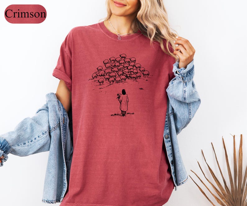 Op de afbeelding: Karmizijnrood T-shirt met een zwarte illustratie van een persoon en een kudde schapen. De persoon houdt een schaap vast. Het shirt heeft korte mouwen en een relaxte pasvorm. Het woord "Crimson" staat in een bordeauxrode rechthoek linksboven.
