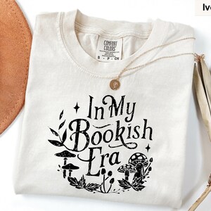 May include: Ivory-colored t-shirt with the text "In My Bookish Era" in black, surrounded by a floral and mushroom design. The shirt has a crew neck and short sleeves. A gold necklace is draped across the shirt.