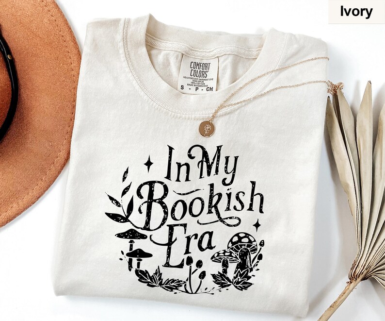 May include: Ivory-colored t-shirt with the text "In My Bookish Era" in black, surrounded by a floral and mushroom design. The shirt has a crew neck and short sleeves. A gold necklace is draped across the shirt.