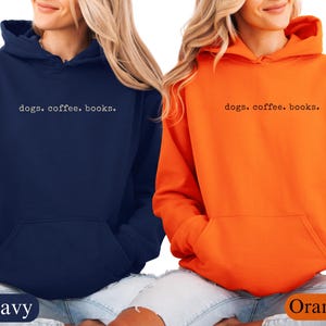 May include: Two hooded sweatshirts, one navy blue and one orange, are displayed. Each sweatshirt has a front pocket and the phrase "dogs. coffee. books." printed in a simple font across the chest. The navy sweatshirt is labeled "Navy" and the orange sweatshirt is labeled "Orange".