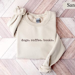 May include: Sand-colored sweatshirt with the text "dogs. coffee. books." in a dark brown font. The sweatshirt has a crew neck and ribbed cuffs and hem. The sleeves are folded up. The background is a light brown wooden surface.