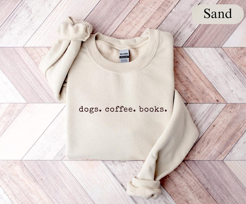 May include: Sand-colored sweatshirt with the text "dogs. coffee. books." in a dark brown font. The sweatshirt has a crew neck and ribbed cuffs and hem. The sleeves are folded up. The background is a light brown wooden surface.