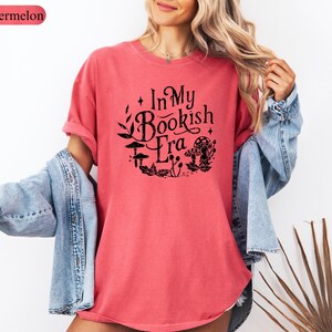 May include: A watermelon-colored t-shirt with the text "In My Bookish Era" in a black, circular design featuring mushrooms and leaves. The shirt is paired with a light blue denim jacket.