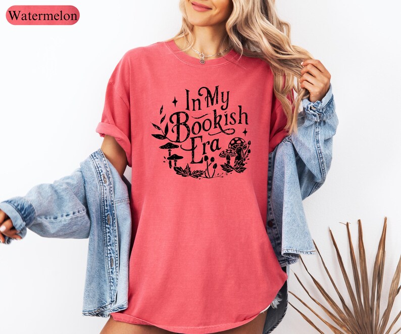 May include: A watermelon-colored t-shirt with the text "In My Bookish Era" in a black, circular design featuring mushrooms and leaves. The shirt is paired with a light blue denim jacket.