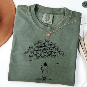 Op de afbeelding: Mosgroen T-shirt met een ronde hals en korte mouwen. Het shirt heeft een zwarte afbeelding van een herder met een schaap en een kudde schapen. Een gouden ketting met een hanger ligt op het shirt.