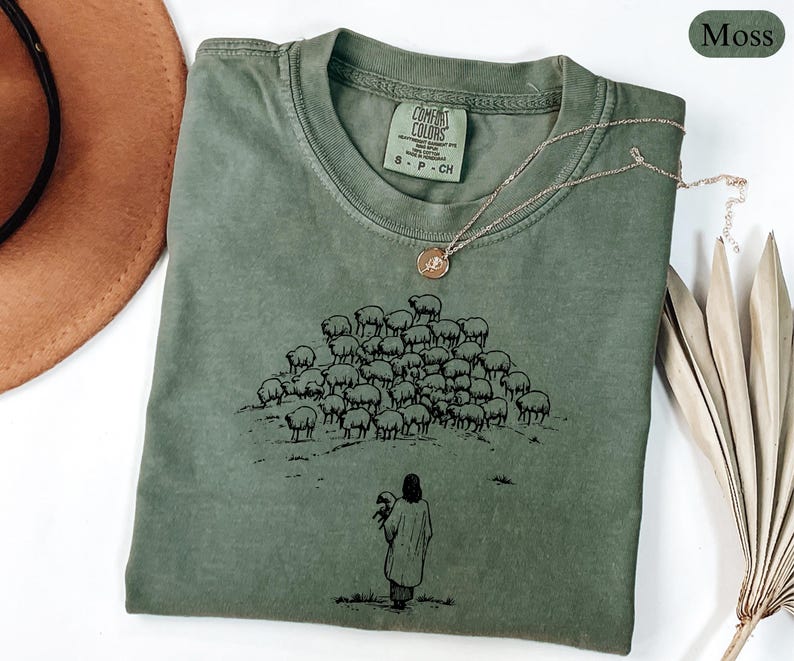 Op de afbeelding: Mosgroen T-shirt met een ronde hals en korte mouwen. Het shirt heeft een zwarte afbeelding van een herder met een schaap en een kudde schapen. Een gouden ketting met een hanger ligt op het shirt.